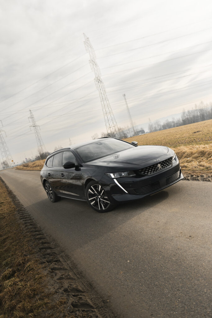 Czarny Peugeot 508 SW pod kątem na drodze, słupy wysokiego napięcia, fotografia.